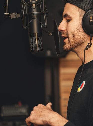 A man wearing headphones and recording vocals in a music studio with a microphone.